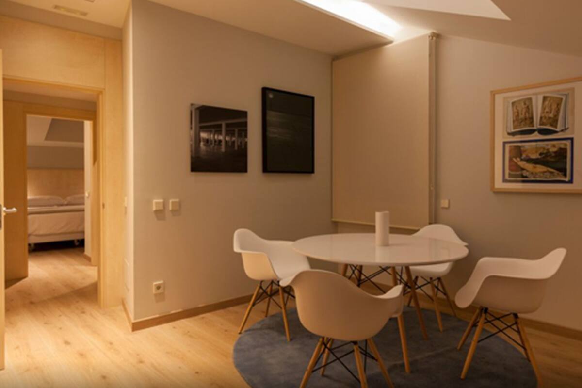 Una habitación moderna con una mesa redonda y cuatro sillas, una cama al fondo y dos cuadros en la pared.