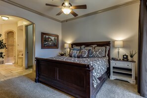 A bedroom with a large bed, a ceiling fan, a nightstand, a lamp, a clock, and a painting on the wall.