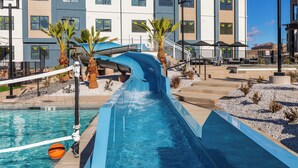 Un toboggan aquatique qui débouche sur une piscine, le tout entouré de bâtiments modernes et de palmiers.