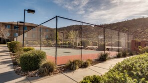 Una cancha de tenis rodeada por una cerca de alambre, abundante vegetación ajardinada y un edificio al fondo.