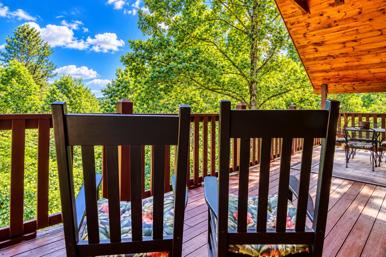 A wooden deck with two chairs and a table, surrounded by lush greenery and a clear sky.