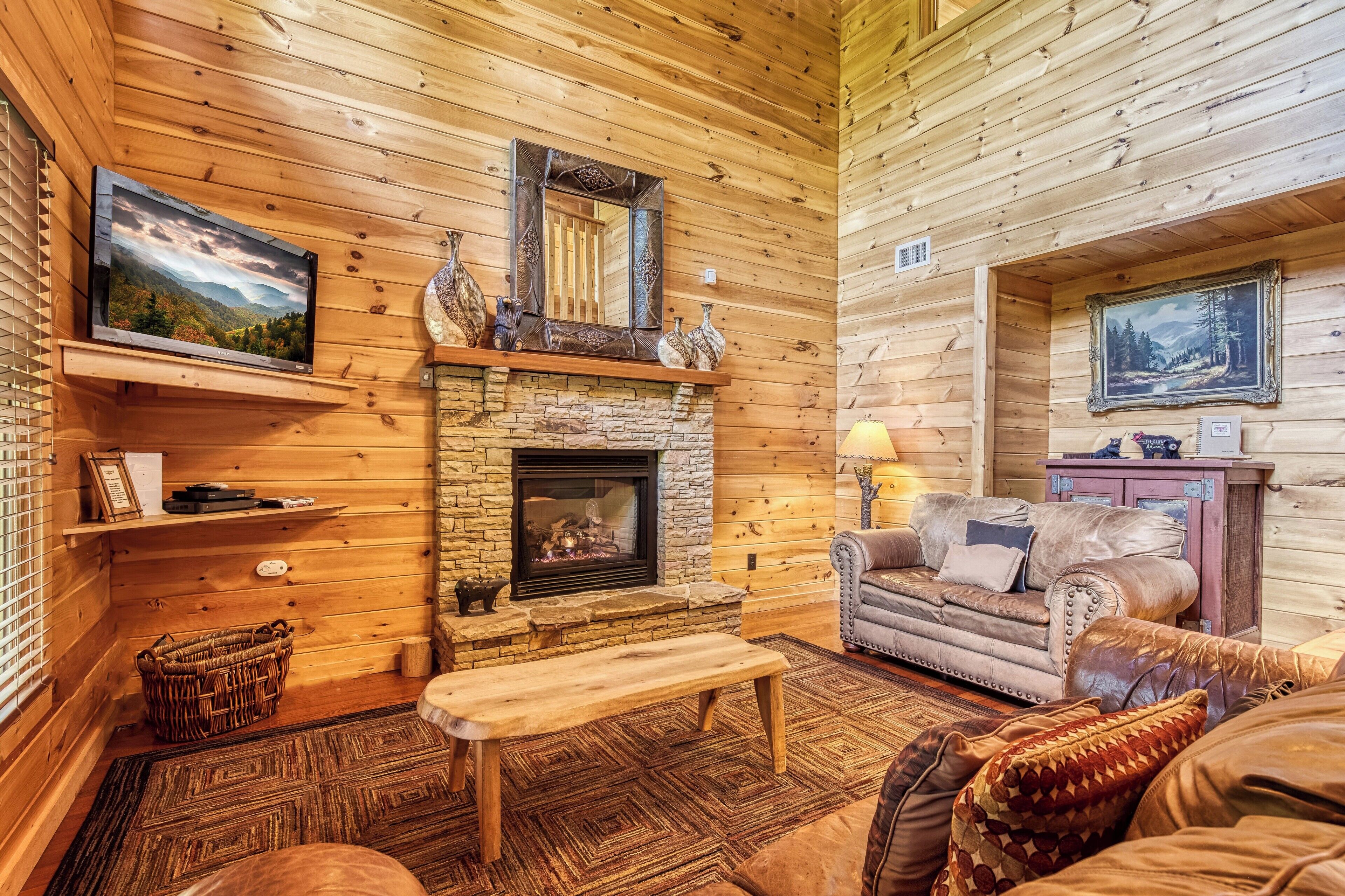 A cozy cabin interior with a stone fireplace, wooden walls, a leather sofa, and a television mounted on the wall.