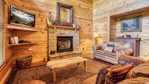 L’intérieur chaleureux d’un chalet avec une cheminée en pierre, des murs en bois, un canapé en cuir et un téléviseur fixé au mur.