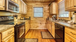 Une cuisine rustique aménagée dans une cabane en rondins, avec des meubles en bois, des appareils électroménagers en acier inoxydable et une fenêtre équipée de stores.