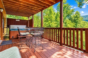 A wooden deck with a hot tub, outdoor seating, and a view of a forested hillside.