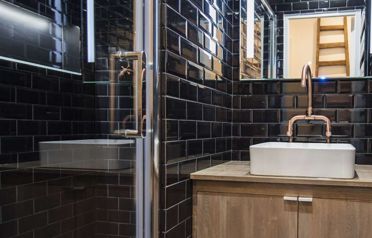 Ein modernes Badezimmer mit schwarzen Fliesenwänden, einem weißen Waschbecken mit Kupferarmaturen und einem Holz-Waschtisch.
