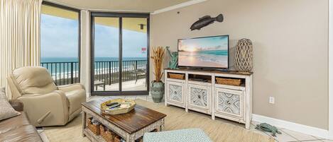 A living room with a sofa, a coffee table, a TV, and a balcony with a view of the beach.