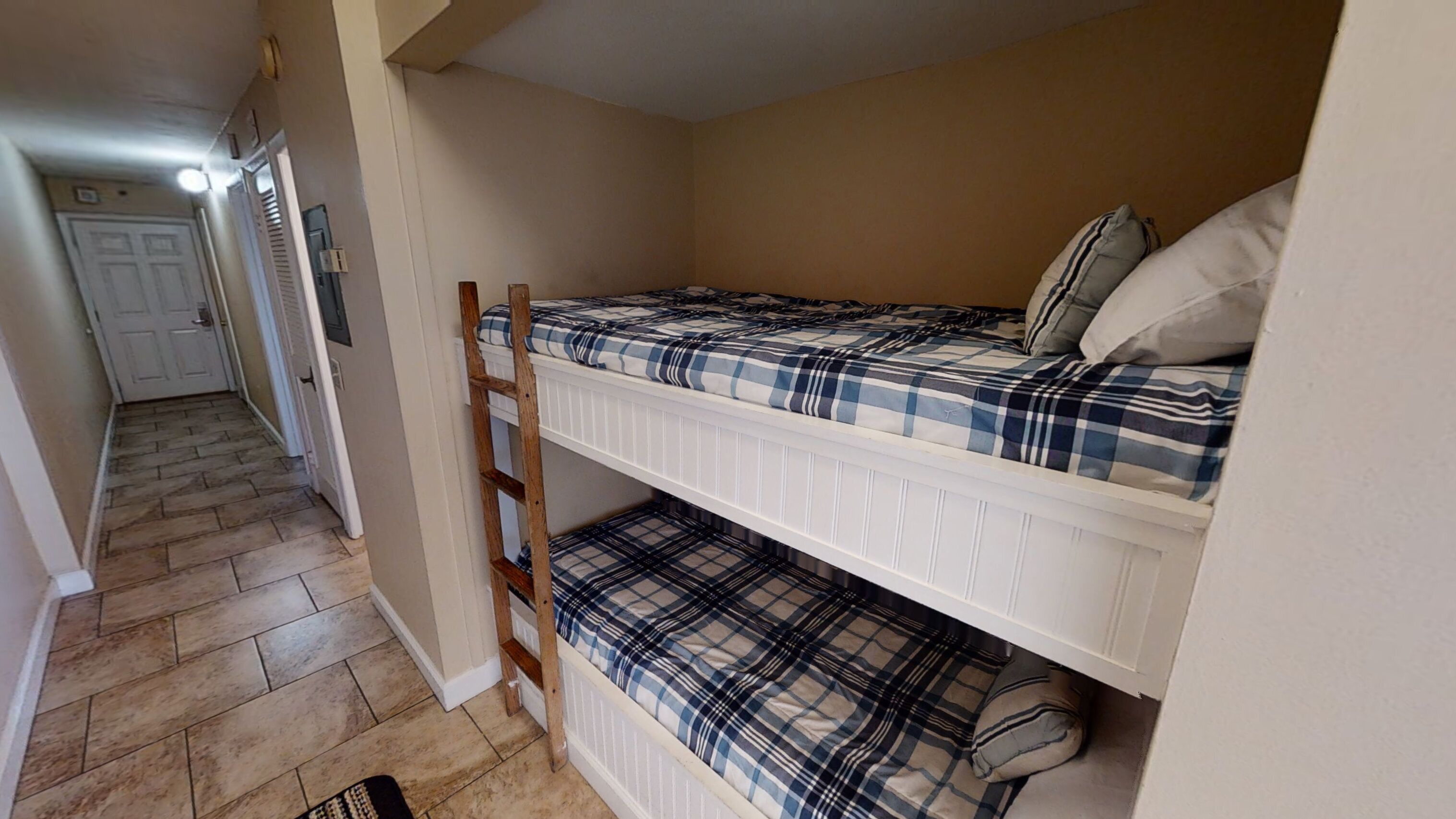 Uma cama beliche com colcha xadrez e travesseiros, uma escada de madeira, e um corredor que dá para uma porta.