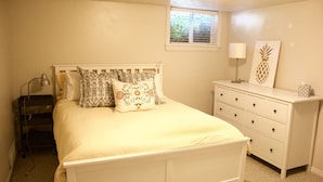 Une chambre à coucher avec un lit blanc, des coussins à motifs, une lampe de chevet, une commode et une fenêtre avec des stores.