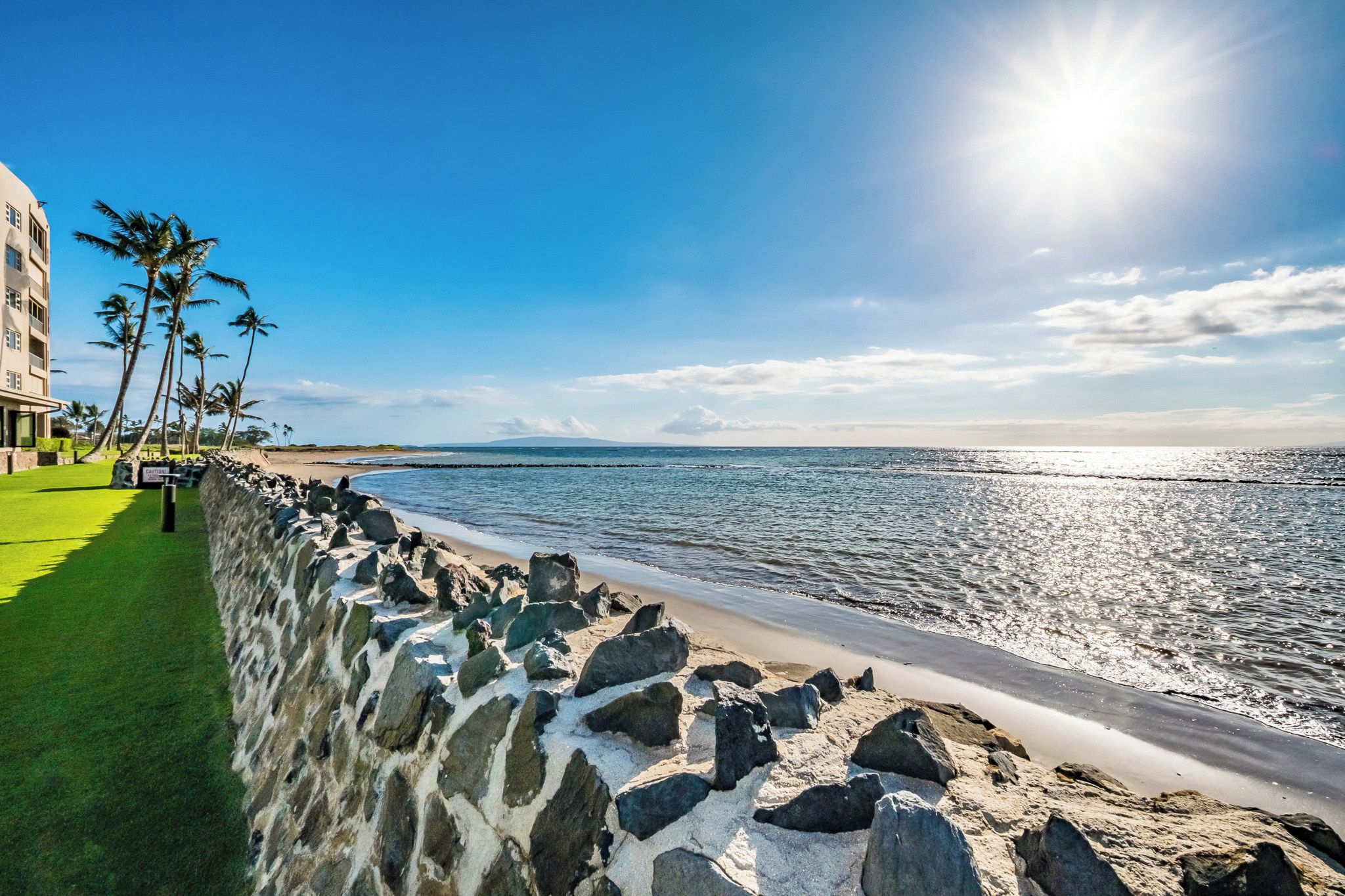 Een hotel aan het strand met een stenen muur, palmbomen en een helder uitzicht op de oceaan.