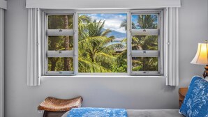 A room with a bed, a window with a view of palm trees and a mountain, a lamp, and a small table with a woven stool. - Menehune Shores #401 4th floor Ocean View, A/C throughout, Great Rates (Kihei)
