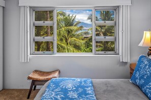 Una stanza con un letto, una finestra vista palme e montagna, una lampada e un piccolo tavolo con uno sgabello intrecciato.