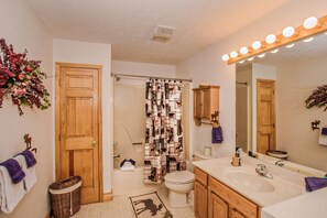 Ein Badezimmer mit Holztür, einer Duschvorhang mit Tiermotiven, einer Toilette, einem Waschbecken mit Spiegel darüber und einem Korb auf dem Boden.