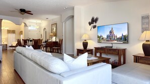 Un salon spacieux avec un grand canapé blanc, un téléviseur diffusant une image de château et un coin repas doté de meubles en bois.