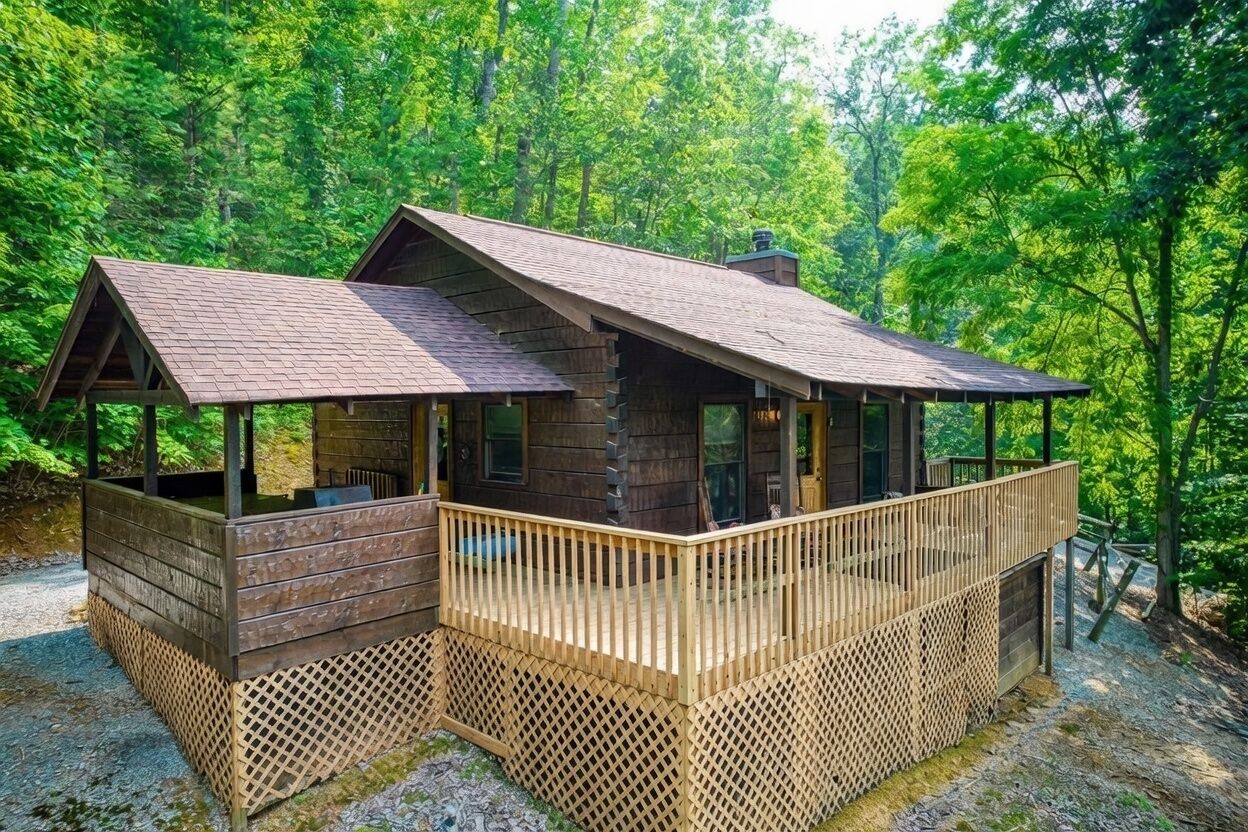 A cabin with a wooden deck and a covered porch, surrounded by dense forest.