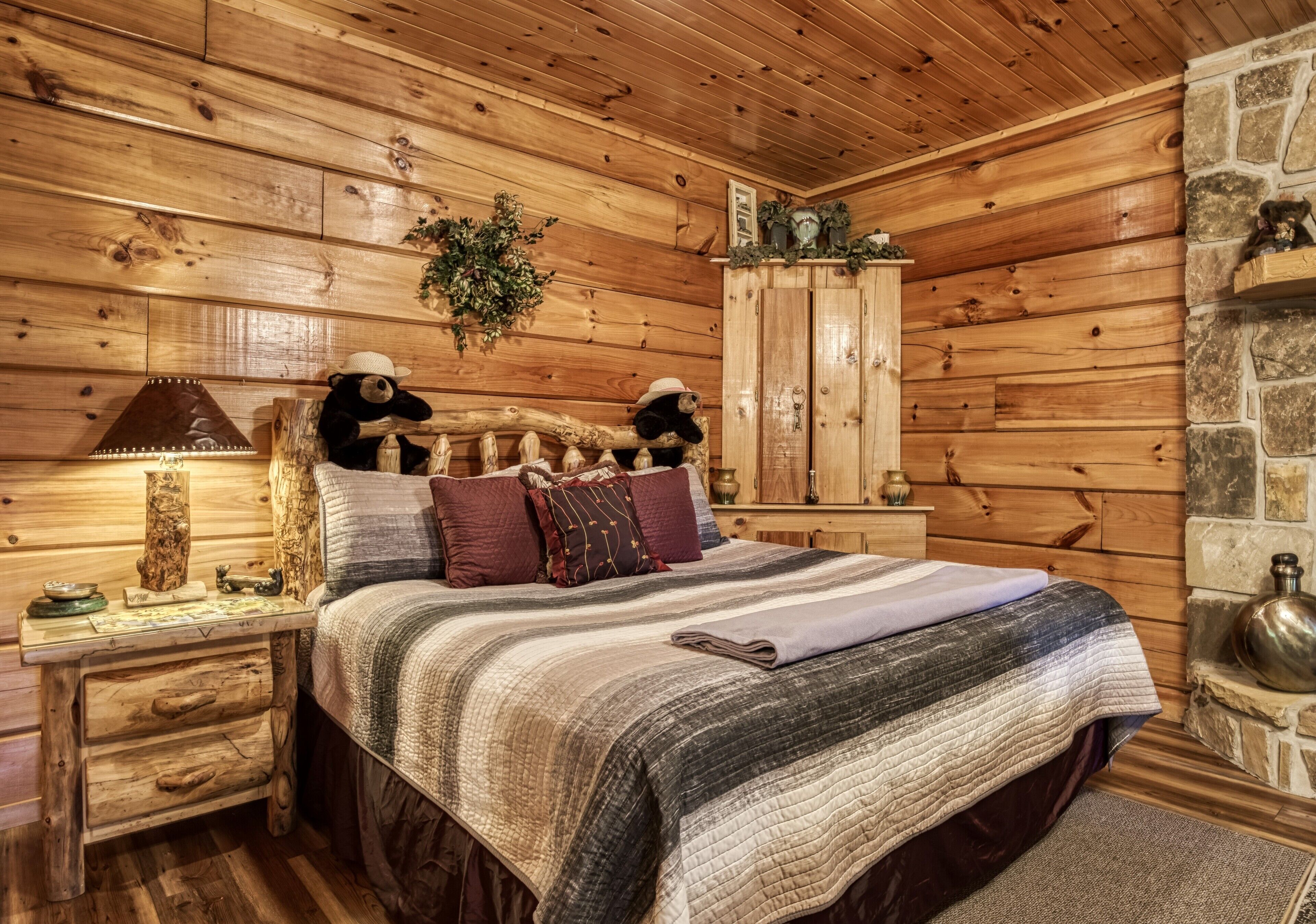 A cozy log cabin bedroom with a wooden bed, a nightstand with a lamp, and a stone fireplace.