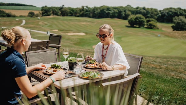 Zwei Personen essen an einem Tisch im Freien mit Blick auf einen Golfplatz.