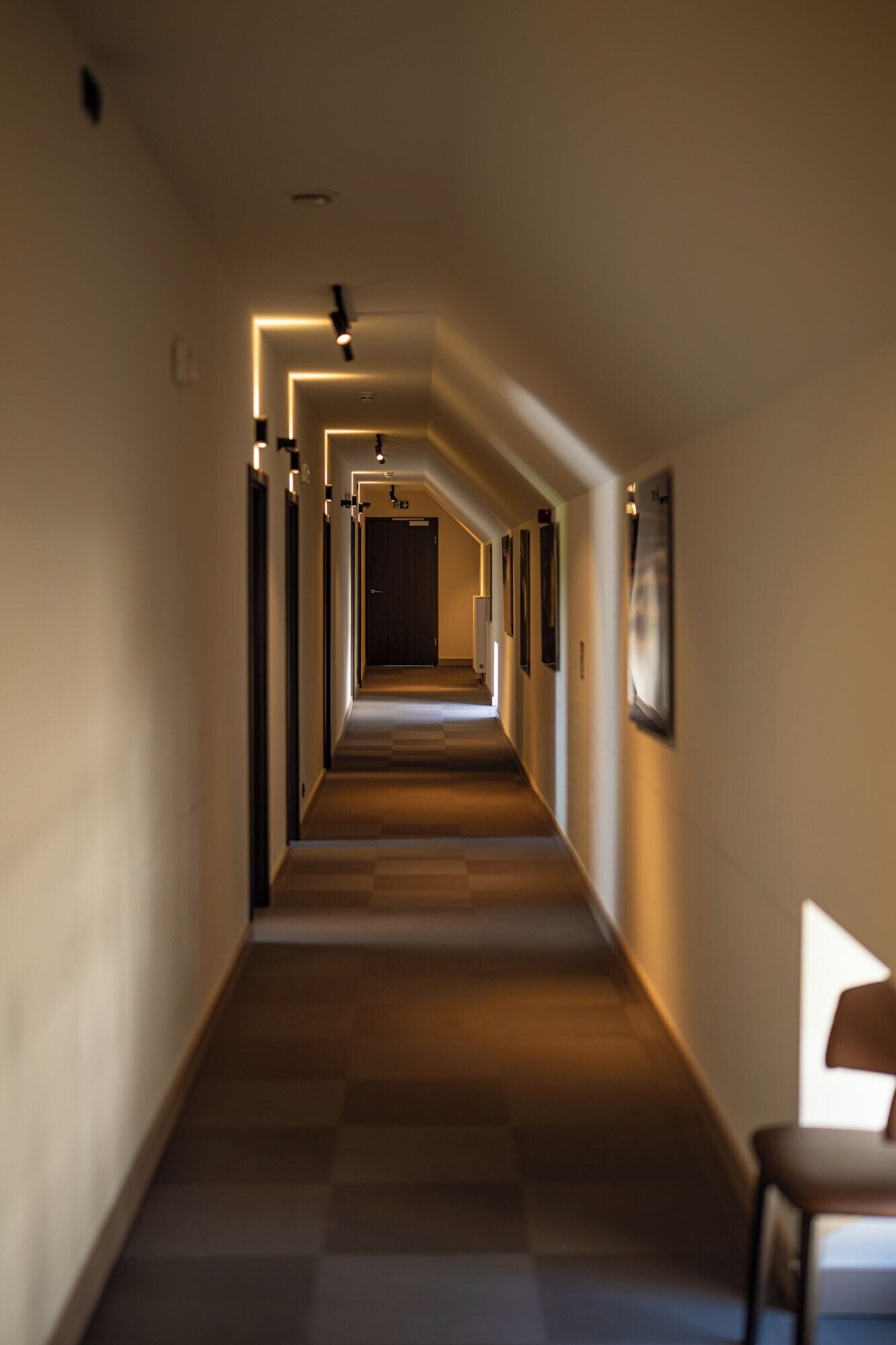 Een lange, smalle hotelgang met een zwart-wit geruite vloer, inbouwverlichting en ingelijste foto's aan de wanden.