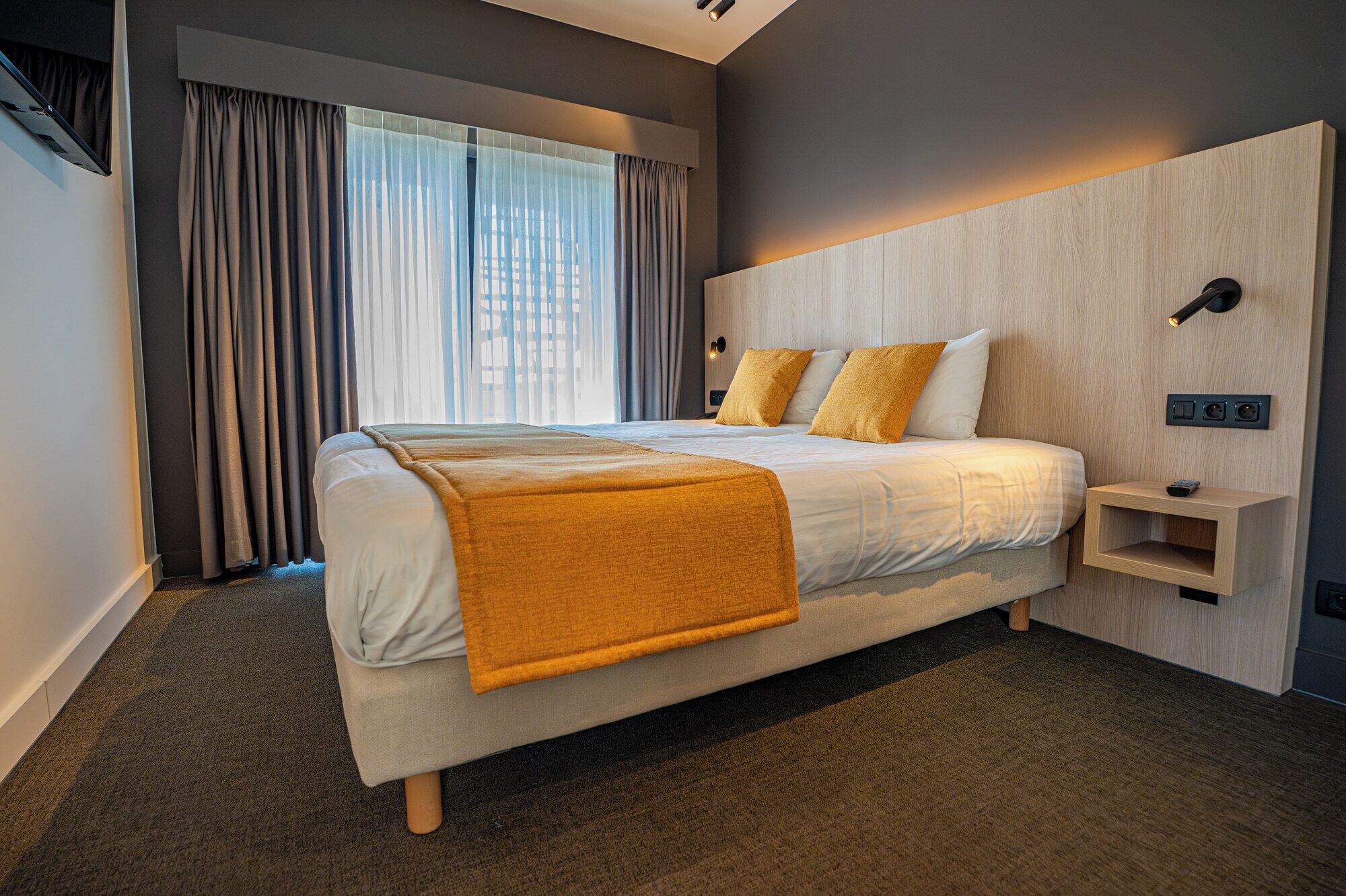 A modern hotel room with a large bed, a bedside table, a wall-mounted TV, and a window with curtains.