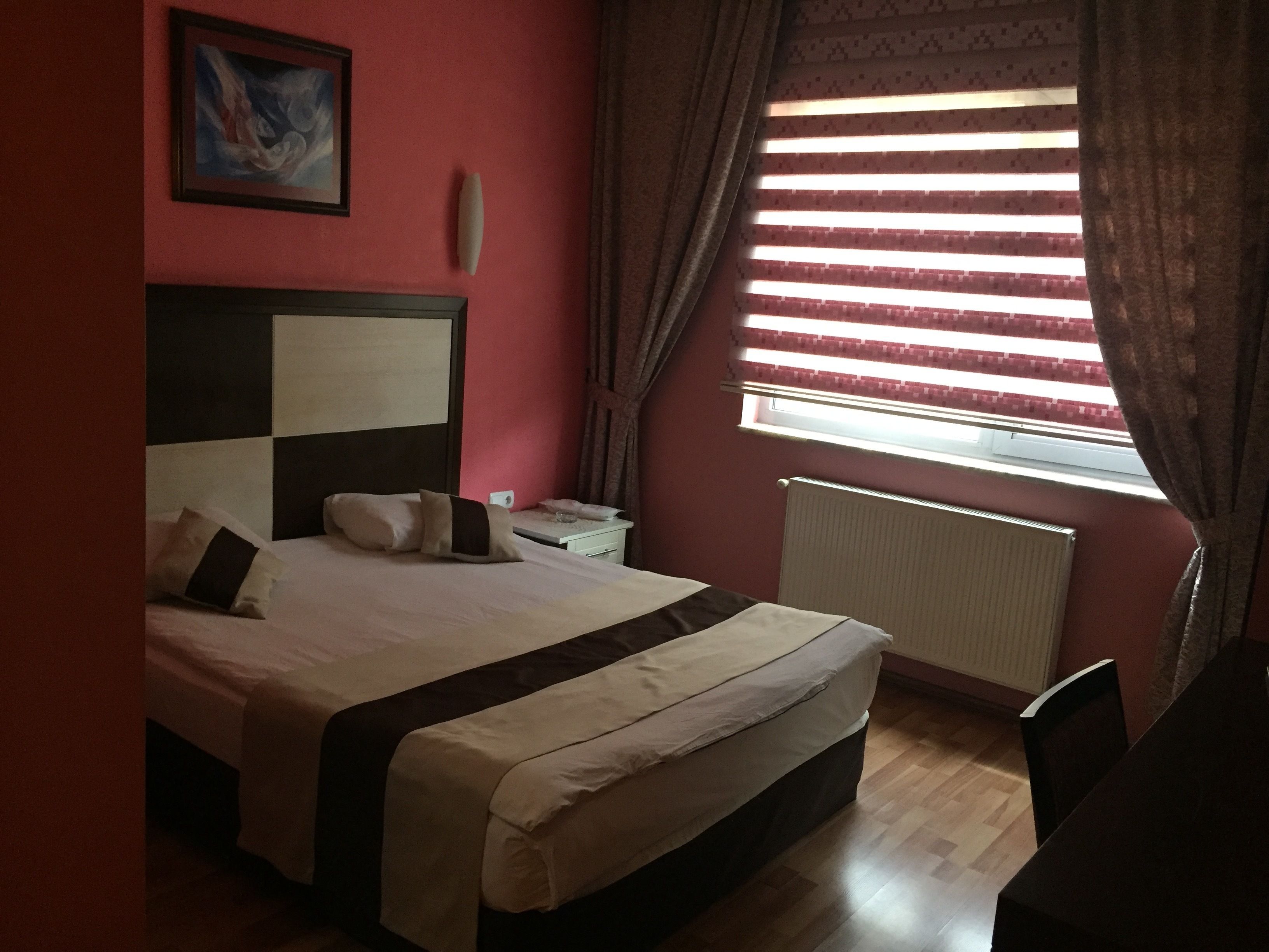 A bedroom with a bed, a nightstand, a painting, a window with curtains, and a radiator.