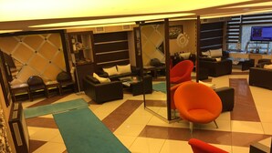 Un área de descanso moderna con piso a cuadros, diversas opciones de asientos que incluyen sofás y sillones, y una pantalla de televisión mostrando contenido.