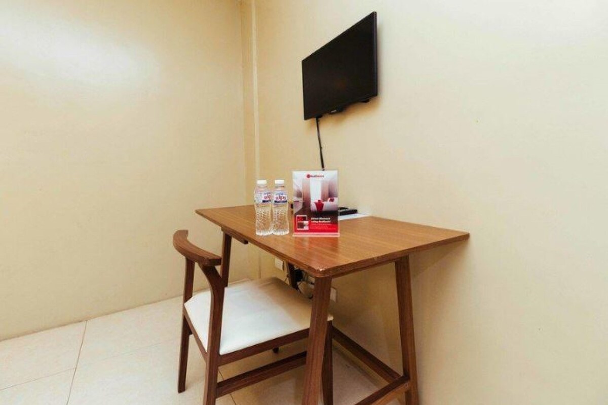 A small wooden table with a white cushioned chair, a flat-screen TV mounted on the wall, and a box of bottled water and a red item on the table.