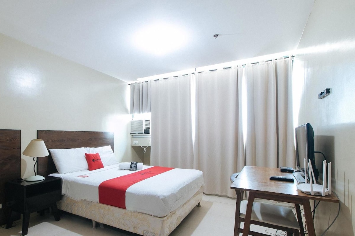 A hotel room with a bed, bedside table, lamp, and a desk with a computer.