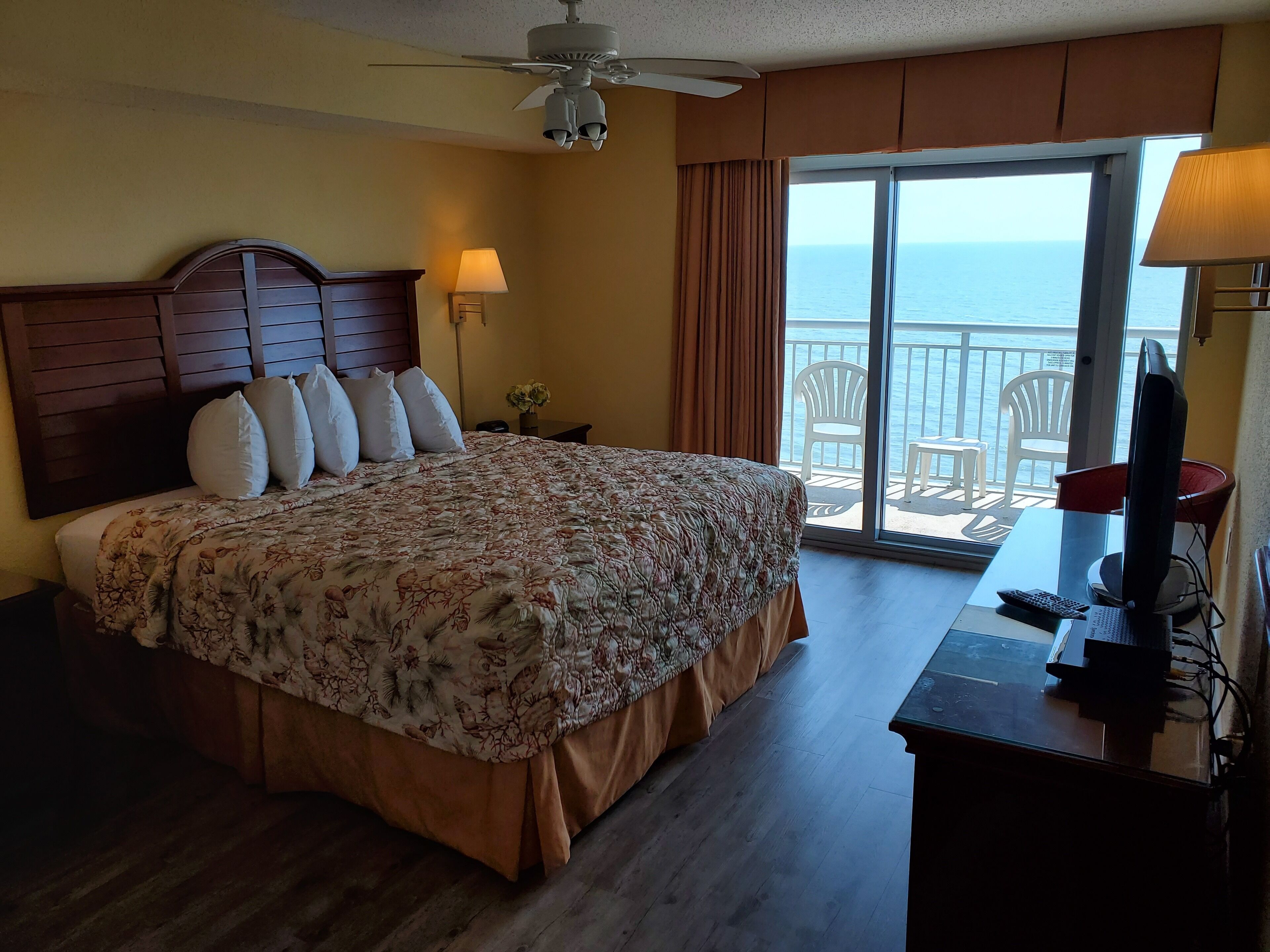 A hotel room with a large bed, a ceiling fan, a desk with a computer, and a view of the ocean through a sliding glass door.