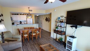 A living room with a sofa, a coffee table, a dining table with chairs, a kitchen area with cabinets and a refrigerator, and a television mounted on the wall. - Stunning 3 BR, 3 Bath Grand Atlantic Direct Oceanfront (Myrtle Beach)