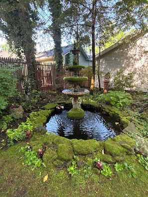 A multi-tiered fountain surrounded by a moss-covered pond, with a wooden fence and a house in the background.