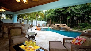 Un espace aménagé au bord de la piscine, avec des meubles en osier, une table en verre sur laquelle sont disposées des boissons, et une vue sur un paysage naturel composé d’arbres et d’une cascade.