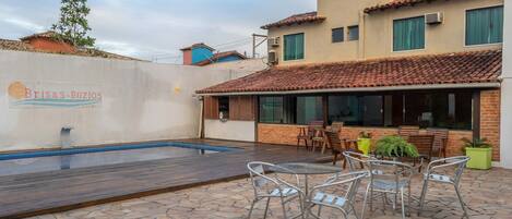 Un área de piscina con una mesa y sillas en la terraza, una piscina y un edificio con tejado de azulejos y ventanas.