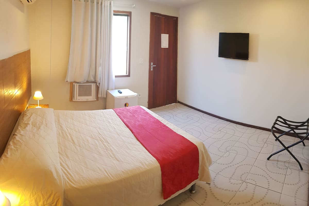 Ett hotellrum med en säng, ett skrivbord, en luftkonditioneringsanläggning, en TV och en stol.
