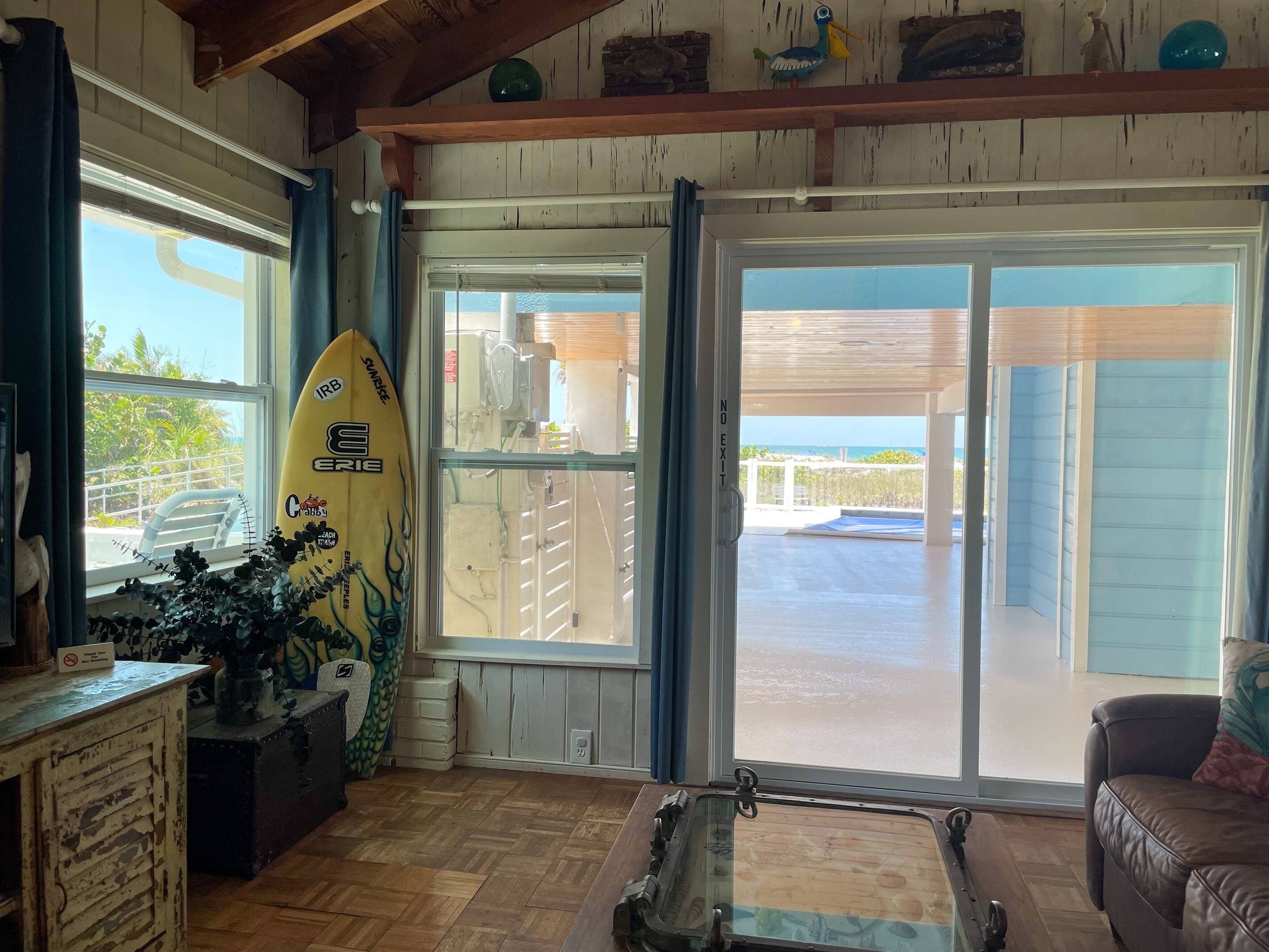 A room with a surfboard, a glass table, a couch, and a view of the beach through a sliding door.