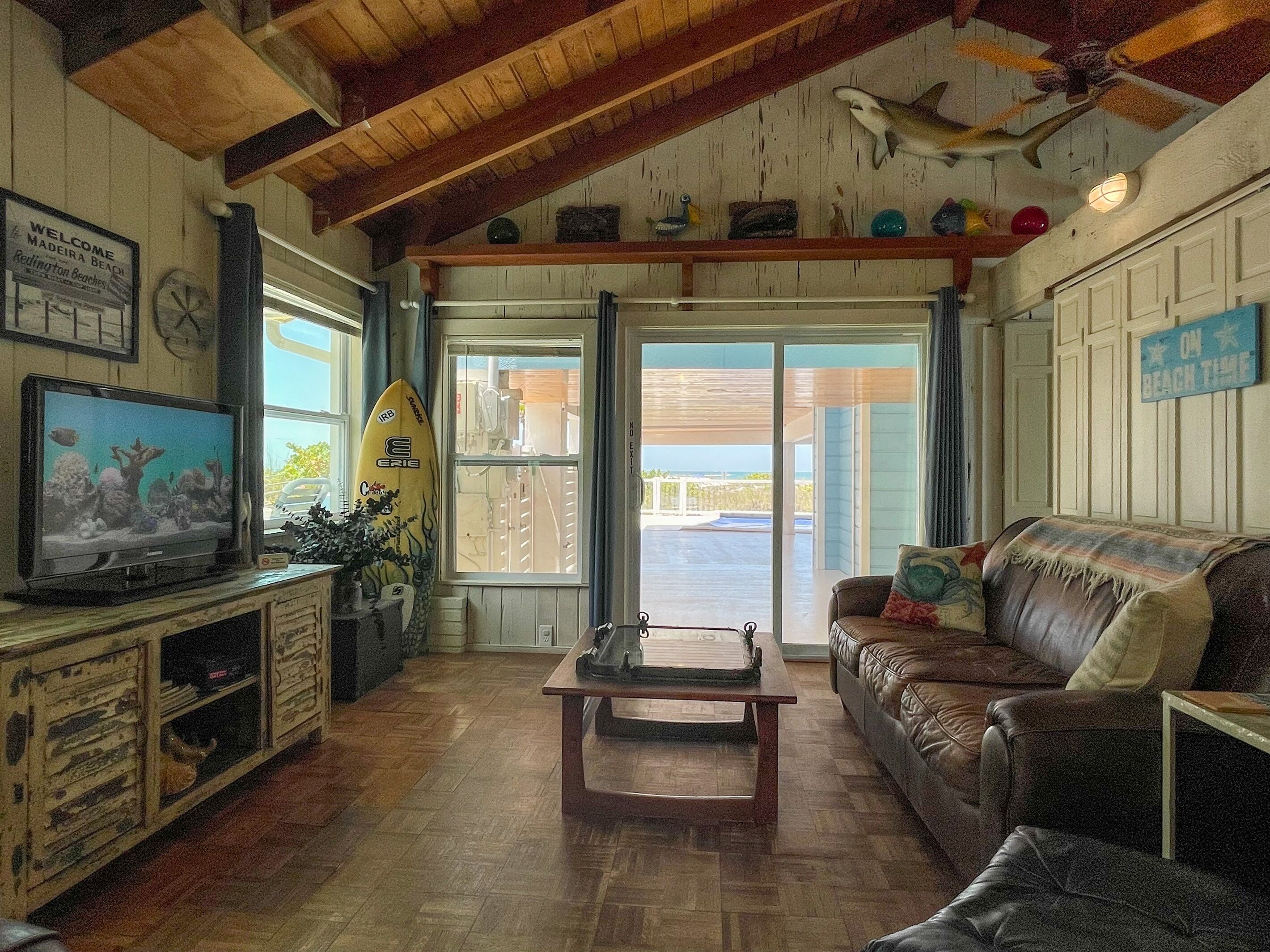 Een gezellige woonkamer met houten plafond, een televisie, een surfplank en uitzicht op het strand door de glazen deur.