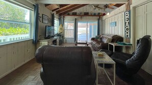 A cozy living room with a leather couch, a coffee table, and a television. A surfboard is visible in the background, and the word "BEACH" is displayed on the wall. - Beach View, Private Plunge Pool, Charming 1938 Bungalow Madeira Beach Gulf-Blvd (Madeira Beach)