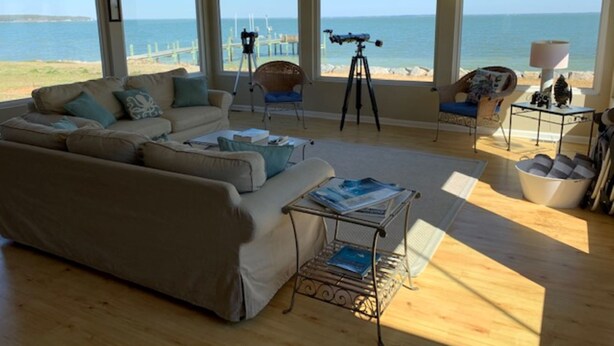 A spacious living room with a large window overlooking the beach, a comfortable sofa, a tripod, and a small table with books.