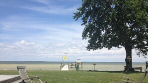 A grassy area with two wooden chairs and a bench, a dock leading to a boat, and a flagpole with a flag.