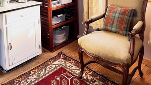 Une cuisine compacte avec un réfrigérateur blanc, un four à micro-ondes et une machine à café sur le plan de travail. Un meuble en bois, doté d’étagères, est adossé au mur, et une chaise à carreaux est placée près d’une fenêtre ornée de rideaux.