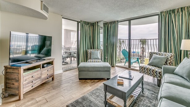 A modern living room with a flat-screen TV, wooden entertainment unit, grey sofa, patterned armchair, and a balcony with green curtains. - 3BR Direct Oceanfront Condo in the Palms (Myrtle Beach)