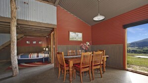 A dining area with a wooden table and chairs, a painting on the wall, and a view of a landscape through a large window. - Studio @ Quins Gap Retreat (Bright)