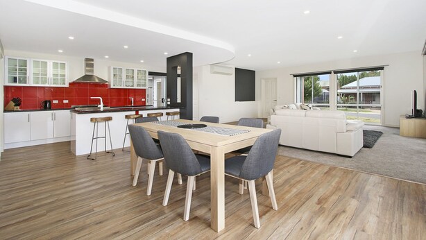 A modern kitchen with red tiles, a dining area with a wooden table and grey chairs, and a white sofa in a living room with large windows. - 3 Peaks Rise - BRIGHT (Bright)