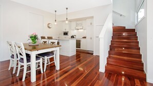 A modern kitchen with white cabinetry, a dining area with a wooden table and white chairs, and a wooden staircase. - Beyond Bliss - Guesthouse 2 (Bright)