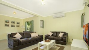A living room with two brown leather sofas, a white coffee table, and a white side table. There are framed pictures on the wall, an air conditioning unit, and a window with green shutters. - River's Reach - Bright (Bright)