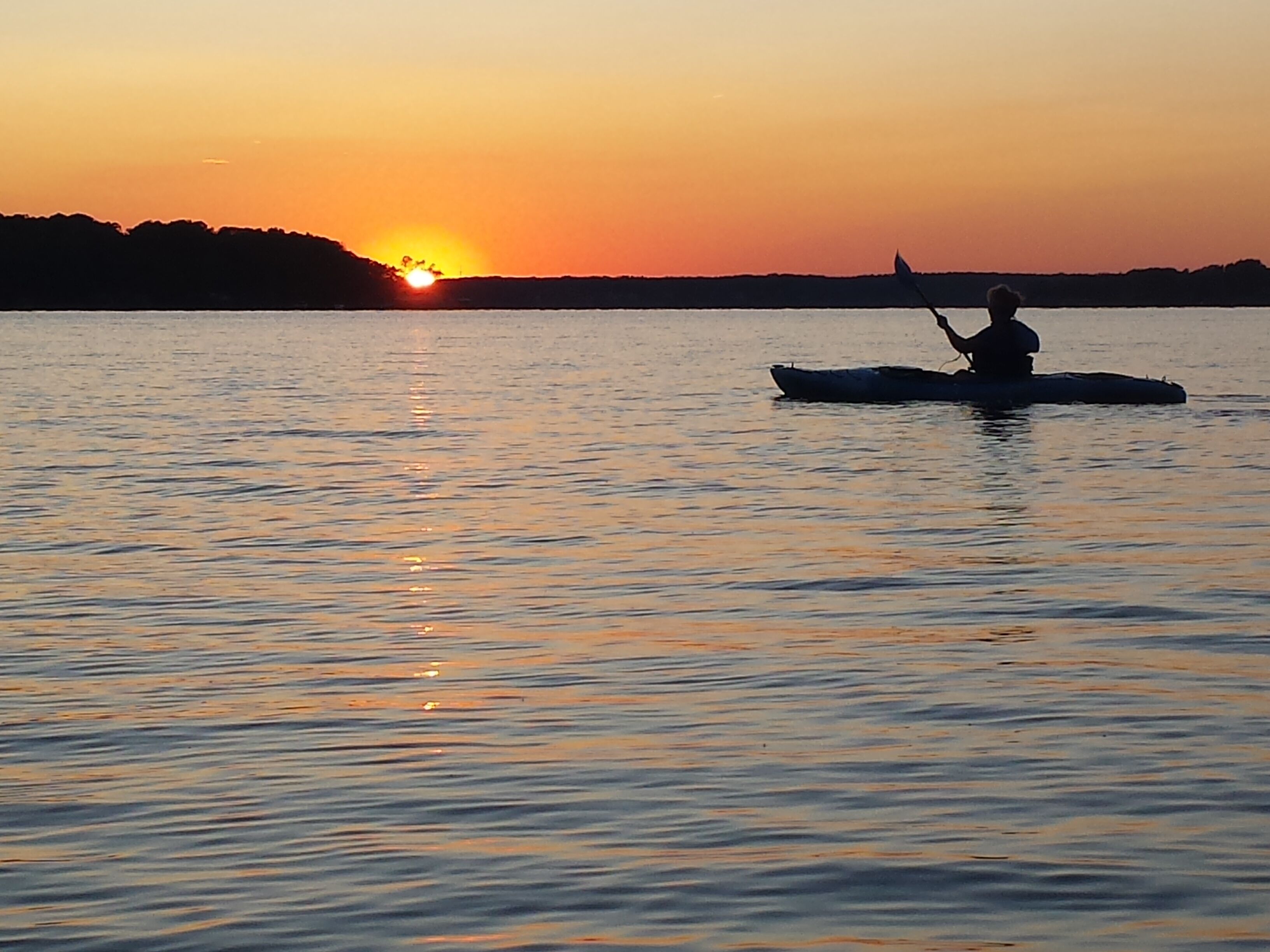 A sunset over a calm body of water with a silhouette of a person paddling a kayak.