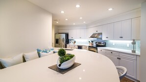 A modern kitchen with white cabinetry, a central island, and a dining area with a white table and blue patterned cushions. - $150 activities - E-gift card when you book - Contemporary Oceanfront Luxury | M2003 (Myrtle Beach)