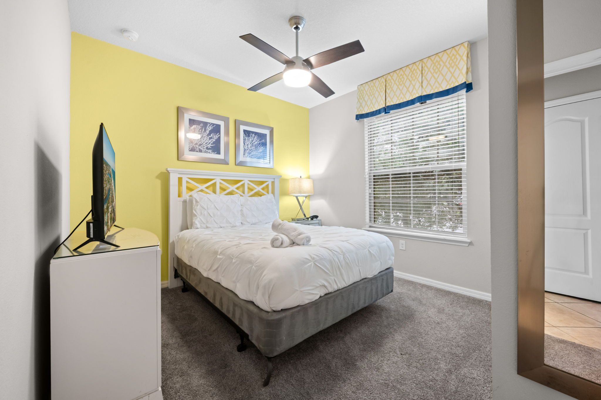 A bedroom with a bed, a ceiling fan, a window with blinds, and two framed pictures on the wall.