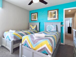 A bedroom with two beds, each with a patterned bedspread and pillows, a ceiling fan, and a door leading to another room.