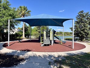 A playground with a blue canopy, red rubber flooring, and a water feature in the background.