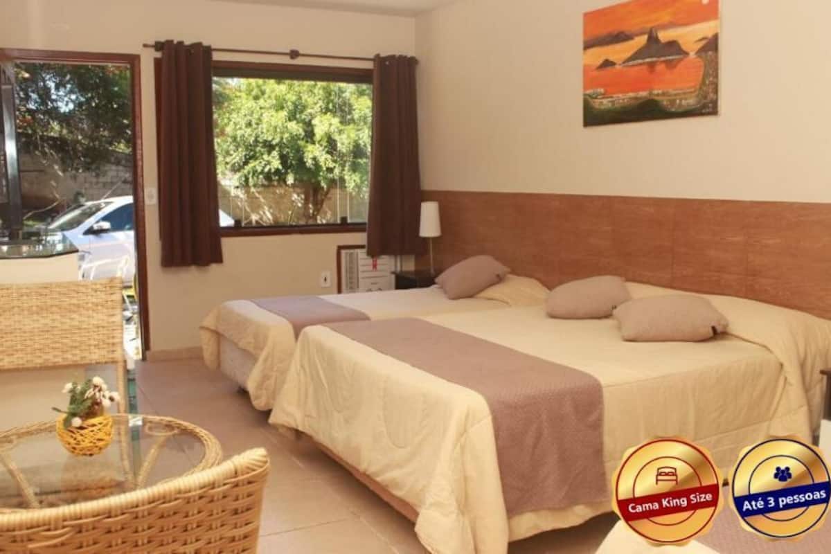 A hotel room with two beds, a wicker chair, a glass table with a basket, and a view of trees and a car outside.